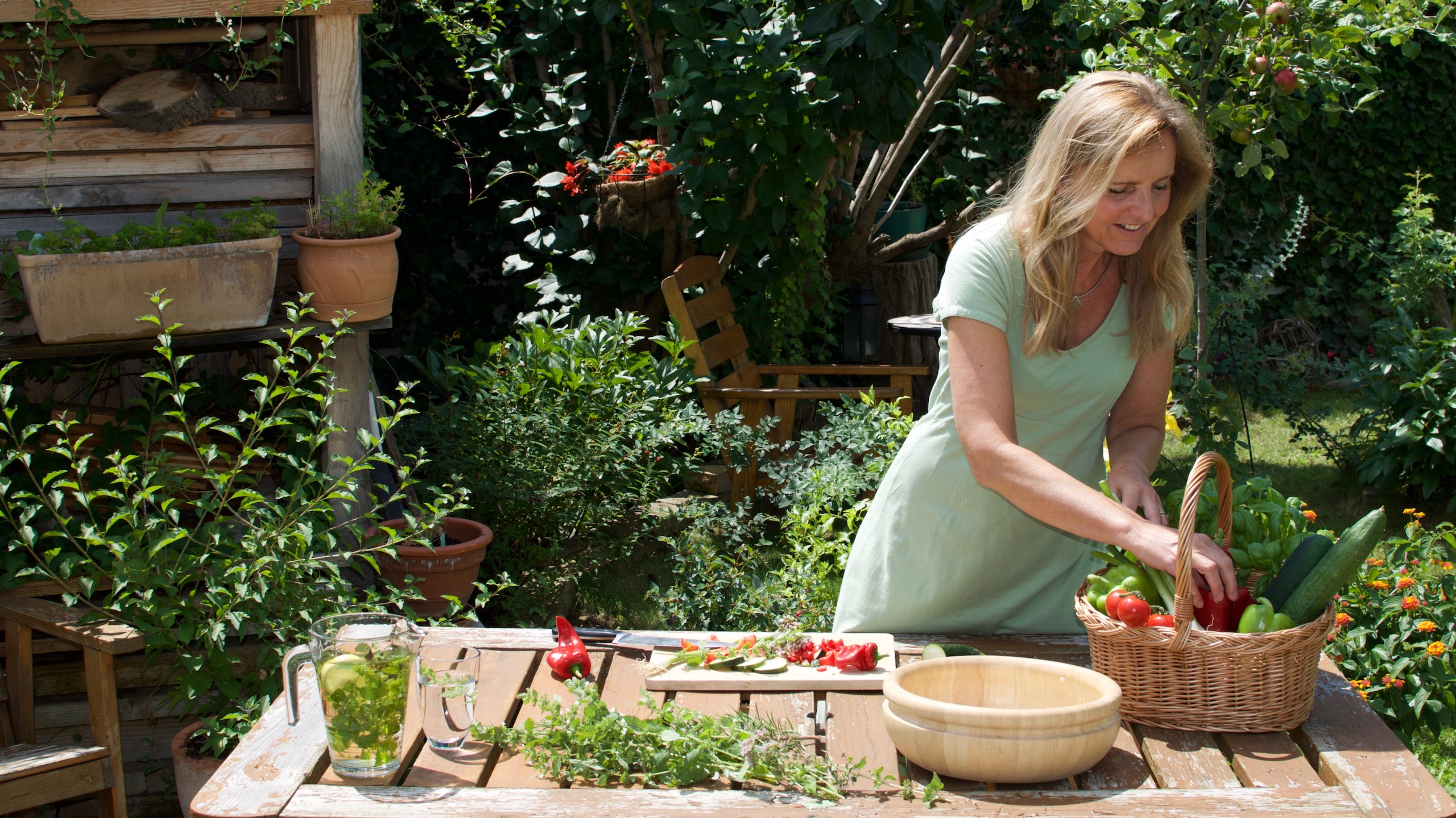 Gaby kocht in ihrem Garten auf einem Holztisch. Sie nimmt gerade etwas aus dem Gemüsekorb.